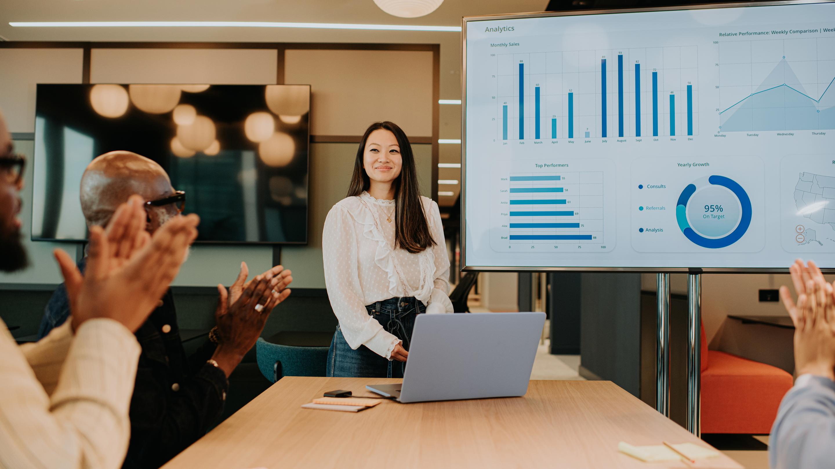 A meeting takes place in a modern office environment. A young woman has completed a successful presentation using a laptop and a huge computer monitor / television. Her colleagues giver her a round of applause for a job well done.